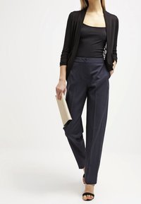 Svart långärmad cardigan över en åtsittande svart topp, i kombination med högmidjade marinblå byxor och svarta sandaletter med remmar; hållande en beige kuvertväska.