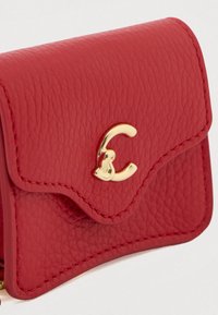 Červená kožená peněženka s texturovaným povrchem, zaobleným klopou a zlatým kovovým logem. Detailní prošití podél okrajů.