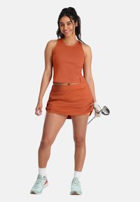 Oranje sportieve tanktop en bijpassende rok, met een getailleerd ontwerp en geplooide zijkanten. Witte sokken en mintgroene sneakers met een golfclub.