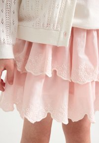 Cardigan bianco lavorato a maglia sovrapposto a una gonna rosa a strati con bordi scallop e ricami floreali bianchi. Sono visibili materiali testurizzati.