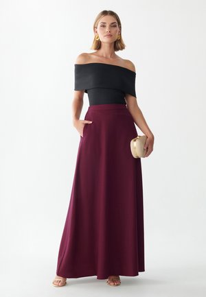 Kvinde står med fronten frem, iført sort off-shoulder top, lang bordeaux nederdel, guldfarvede øreringe, holder en lille guldfarvet clutch og sandaler.