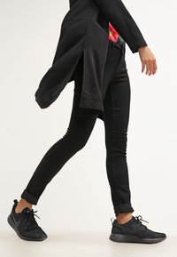 Jeans negros ajustados combinados con una camiseta de manga larga negra. Los zapatos son zapatillas deportivas negras con un diseño texturizado, con los cordones visibles.
