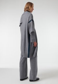 Cárdigan largo de punto gris con aberturas laterales y franjas de acento azul, combinado con pantalones de pierna ancha grises a juego y calzado negro.