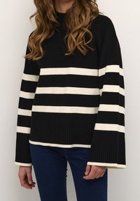 Sweter w czarno-białe paski o prążkowanej fakturze, z wysokim kołnierzem, szerokimi rękawami i kontrastowymi poziomymi liniami. Połączony z niebieskimi dżinsami.