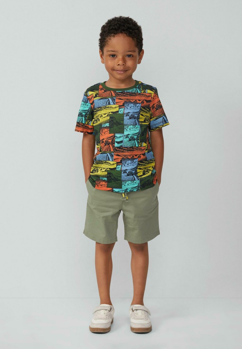 Kleurvol T-shirt met korte mouwen met een collage van landschappen in oranje, groen, blauw en geel. Gepaard met lichtgroene shorts en witte schoenen.