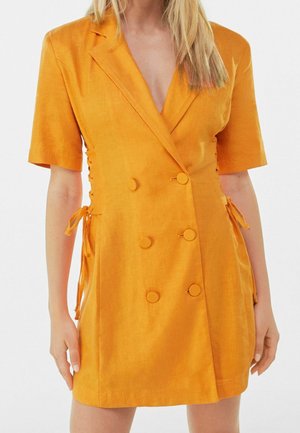 Femme portant une robe blazer orange à manches courtes et boutonnée doublement, avec des détails noués sur les côtés, debout contre un fond blanc uni.