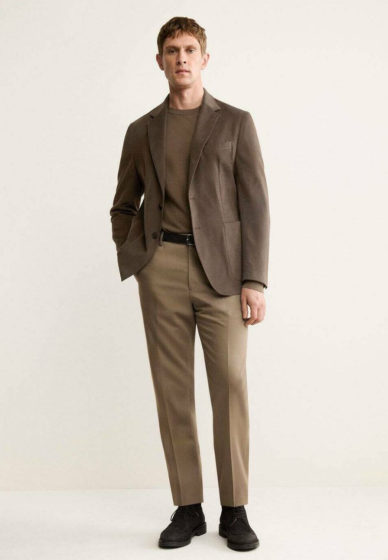 Brown tailored blazer over a brown sweater, paired with beige trousers. Accented with a black belt and dark shoes. Simple, modern design.