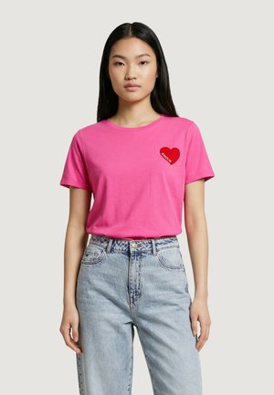 Donna che indossa una maglietta rosa intenso con un piccolo cuore rosso e la parola "AMOUR" sul petto, abbinata a jeans a vita alta di colore azzurro chiaro.