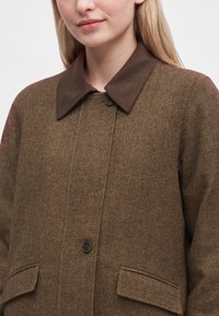 Femme portant un manteau marron à motif chevrons avec un col marron foncé et un devant boutonné, dévoilant partiellement le haut du torse et le visage.