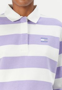 Lilac and white striped polo shirt with a flat collar, featuring a small blue logo patch on the chest. Made of soft, woven fabric.