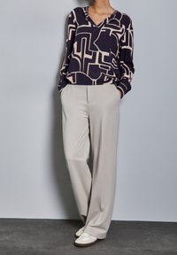 Mörkblå och beige mönstrad långärmad topp med V-ringning, kombinerad med ljusgrå vida byxor och beiga sneakers på en grå bakgrund.