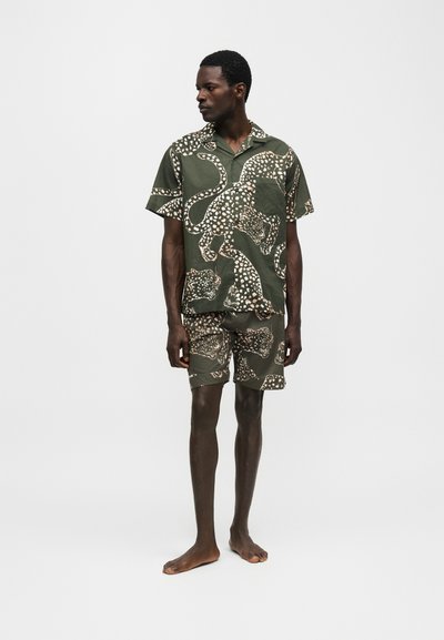Άνδρας που στέκεται ξυπόλητος φορώντας ταιριαστή σκούρα πράσινη κοντομάνικη μπλούζα και σορτς με μεγάλο μπεζ λεοπάρ μοτίβο.