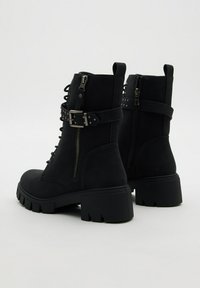 Botines negros de tacón grueso con cremalleras laterales, cordones en la parte delantera y correas con tachuelas en forma de hebilla sobre un fondo claro y liso.