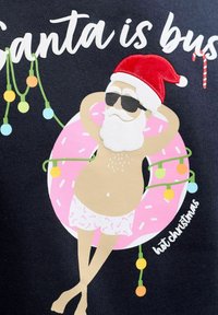 Santa v brýlích a klobouku se povaluje na růžovém nafukovacím donutu, na kterém je omotané vánoční osvětlení, a má na sobě šortky s potiskem cukrové třtiny.