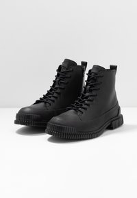 Botas de tobillo negras con cordones y cremalleras a los lados, con suelas de goma texturizadas, exhibidas sobre una superficie blanca con un fondo simple.