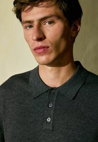 Dunkelgraues Poloshirt mit strukturierter Oberfläche, ausgestattet mit einem klassischen Kragen und drei schwarzen Knöpfen an der Knopfleiste. Nahaufnahme des Ausschnitts.