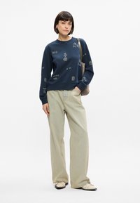 Σκούρο μπλε μπλούζα με λευκά κεντημένα σχέδια, στρογγυλή λαιμόκοψη και μακριά μανίκια, συνδυασμένη με ανοιχτό μπεζ παντελόνι φαρδιάς γραμμής και μαύρα παπούτσια.