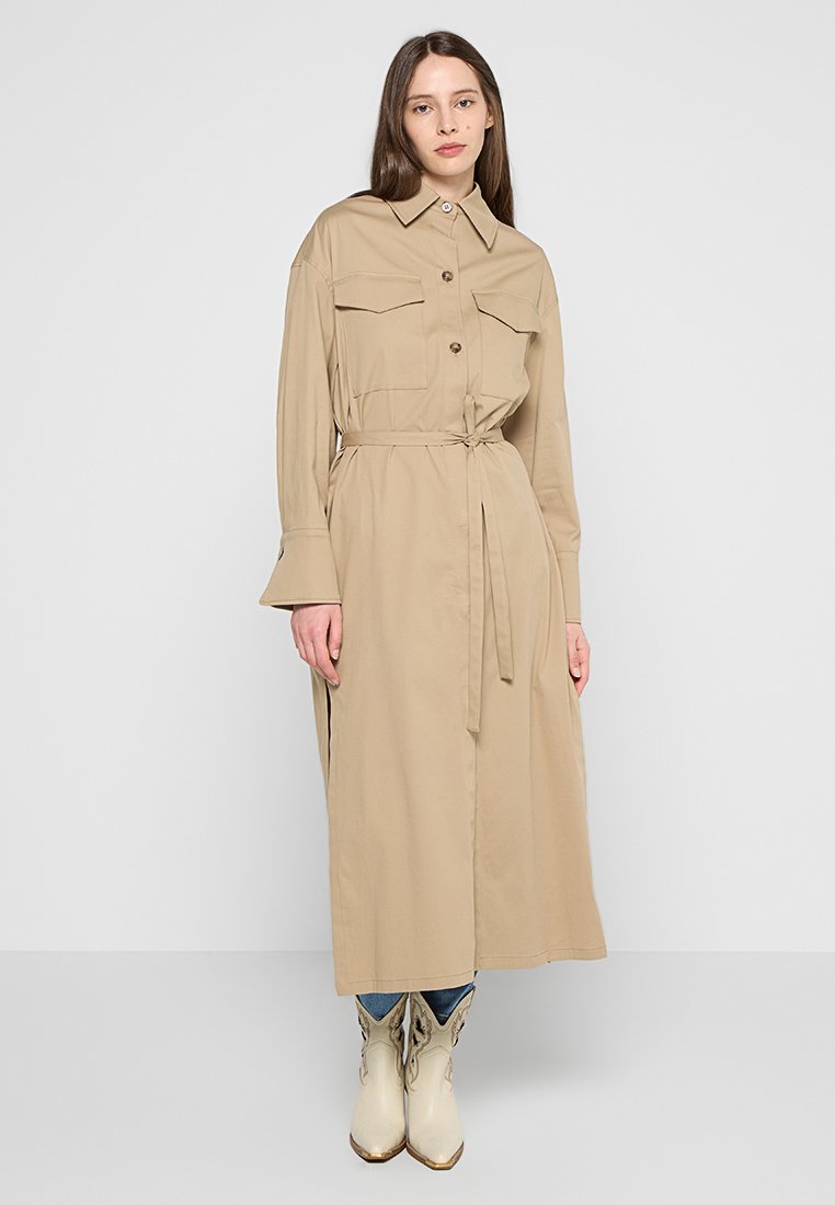 Beżowa sukienka koszulowa z długim rękawem, z dwoma kieszeniami na piersiach, z zapięciem na guziki i paskiem w talii. Posiada rozszerzane mankiety i boczne rozcięcia.