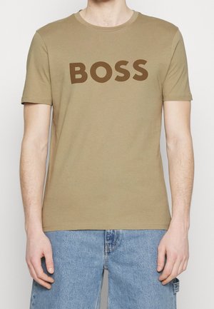 Person trägt ein beige-farbenes Kurzarmhemd mit dem Wort "BOSS" in großen braunen Buchstaben und blaue Denim-Jeans.