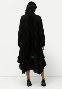 Vestido negro con pliegues verticales, mangas largas, dobladillo ondulado y superposición transparente. Combinado con zapatos de tacón alto.