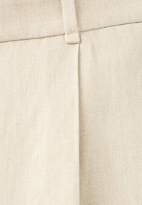Gros plan sur un pantalon en tissu beige montrant une passants de ceinture et un détail de couture centrale.