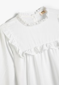 Hvid bluse med frynset halsudskæring og rynkedetaljer langs yoke'en. Stoffet er glat med en let tekstur, der egner sig til lag-på-lag.