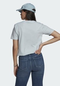 Šviesiai pilka trumpomis rankovėmis marškinėliai su apvalia iškirpte, derinami su tamsiai mėlynomis džinsinėmis kelnėmis, turinčiomis galinius kišenes. Modelis dėvi šviesiai mėlyną kepurę.