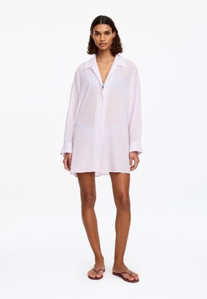 Femme debout portant une chemise oversize rose clair boutonnée comme une robe avec des tongs bordeaux sur fond blanc.