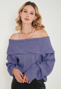 Vijolična svetra off-shoulder z rebrastim teksturo, širokim izrezom in oversized rokavi. Nosi se z složenimi zlatimi verižicami.