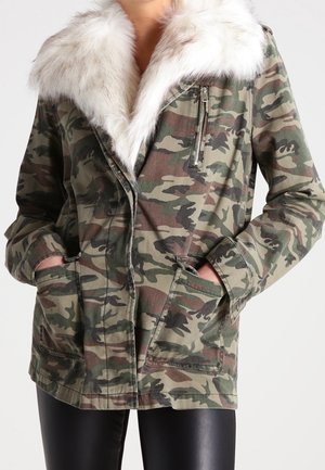 Veste camouflage avec grand col en fourrure blanche, poche poitrine zippée et deux poches frontales inférieures, portée avec un pantalon noir.