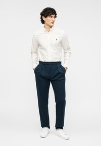 Άνδρας που στέκεται με τα χέρια στις τσέπες, φοράει λευκό πουκάμισο με κουμπιά, μπλε πλισέ παντελόνια και λευκά αθλητικά παπούτσια, κοιτάζοντας προς τα πλευρά.