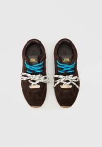 Sapatilhas de camurça castanha com atacadores azul e branco padrão. Detalhes em ouro e logótipo na lingueta. Sola de borracha.