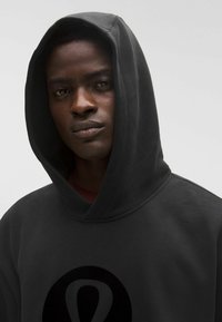 Fekete kapucnis pulóver nagy, kör alakú logóval. Simább, puha anyag, laza fazonnal. A kapucni a vállakra lóg, a nyak szabadon marad.