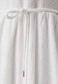 Tissu blanc texturé avec un cordon tressé noué à la taille. Présente un embout métallique sur le cordon pour un détail supplémentaire.