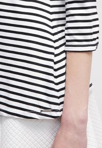 Svart-och-vit randig topp med trekvartsärmar, som har en sydd fåll och en liten metallisk logodetalj på sidan.