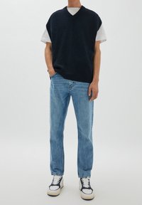 Marinblå stickad ärmlös tröja över en vit t-shirt, i kombination med ljusblå denimjeans och vita sneakers med svarta detaljer.