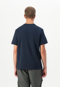 Mann mit kurzen lockigen Haaren, der ein einfaches dunkelblaues T-Shirt und graue Cargo-Hosen trägt, stehend mit dem Rücken zu einer weißen Hintergrund.