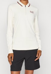 Bílá dlouhá polo košile s předním zipem a límcem se burgundskými pruhy, v kombinaci s černými kraťasy ke kolenům na stojícím člověku.