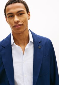 Νεαρός άνδρας με κοντά σγουρά μαλλιά φοράει λευκό πουκάμισο με κουμπιά κάτω από μπλε σακάκι μπροστά σε απλό φωτεινό φόντο.