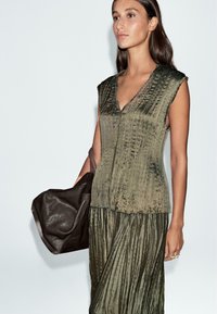 Blusa smanicata verde oliva con texture, scollo a V e dettagli plissettati, abbinata a una borsa in pelle marrone scuro.