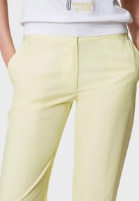 Pantaloni giallo chiaro con una texture liscia, dotati di tasche laterali e una vestibilità su misura. La vita è elasticizzata per maggiore comfort.
