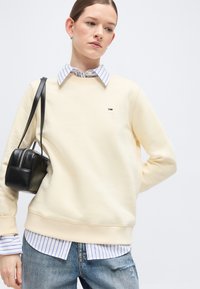 Νεαρή γυναίκα που φοράει μια κρεμ φούτερ πάνω από ένα μπλε-ριγέ πουκάμισο, κρατώντας μια μαύρη δερμάτινη τσάντα ώμου, με σκισμένο μπλε τζιν.