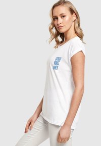 Weißes Baumwoll-T-Shirt mit umgeschlagenen Kurzärmeln, auf der Vorderseite mit blauer Schrift "NUR GUTE VIBES". Glatte Textur und lockere Passform.