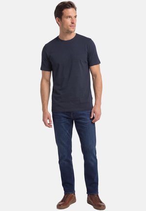 Man staat, draagt een effen donkerblauw T-shirt, blauwe spijkerbroek en bruine leren sneakers, kijkt iets naar rechts.