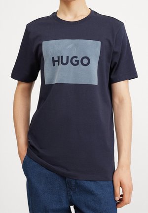 Mand iført en mørkeblå T-shirt med en stor lysegrå firkant og ordet "HUGO" trykt med mørke bogstaver på brystet.