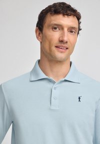 Ljusblå pikétröja med texturerad tyg, med en treknapplist och en liten broderad golfare i marinblått på vänster bröst.