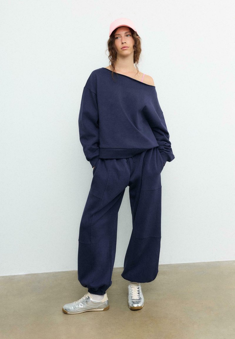 Ung kvinde iført navyblå oversized sweatshirt og bukser, lyserød kasket og sølvfarvede sneakers, stående mod en ensfarvet lys væg og beige gulv.