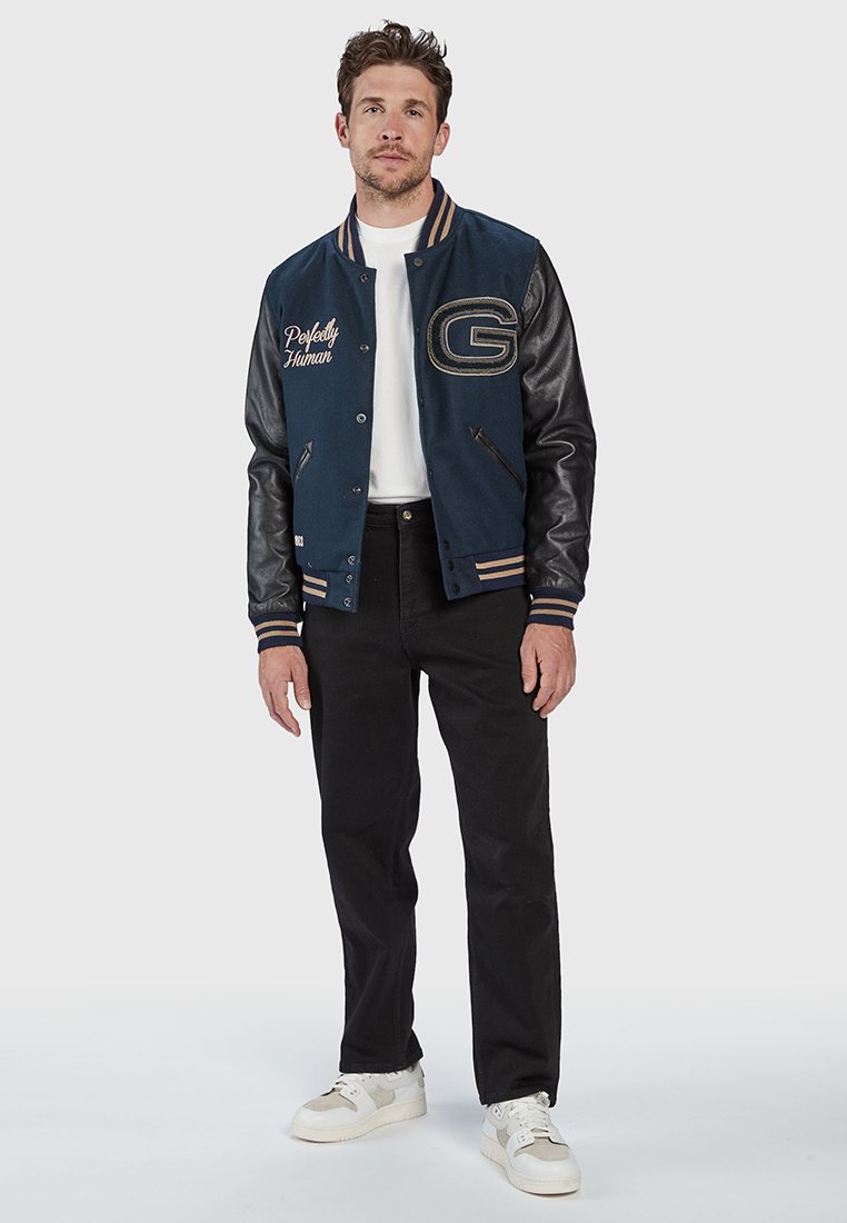 Marineblauwe en zwarte varsityjack met leren mouwen, voorzien van geborduurde tekst "Perfectly Human" en de letter "G," gecombineerd met zwarte broek en witte sneakers.