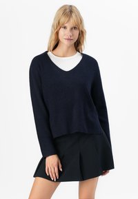 Femeie blondă purtând un pulover bleumarin cu decolteu în V peste un top alb și o fustă plisată neagră, stând cu spatele la un fundal alb simplu.