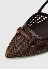 Mocassin tissé marron à bout pointu, avec un accent en cuir foncé et une sangle décorative incurvée avec des éléments métalliques.